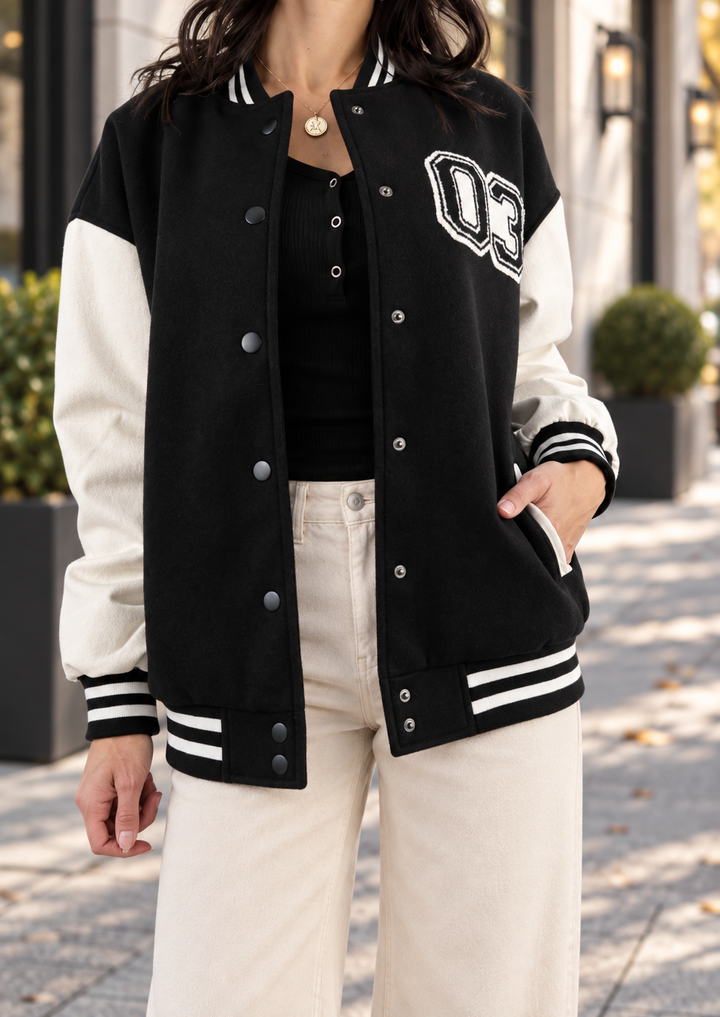 A woman wearing a black and cream varsity jacket with 'DC' lettering on the chest, paired with cream pants, standing outdoors on a sidewalk.