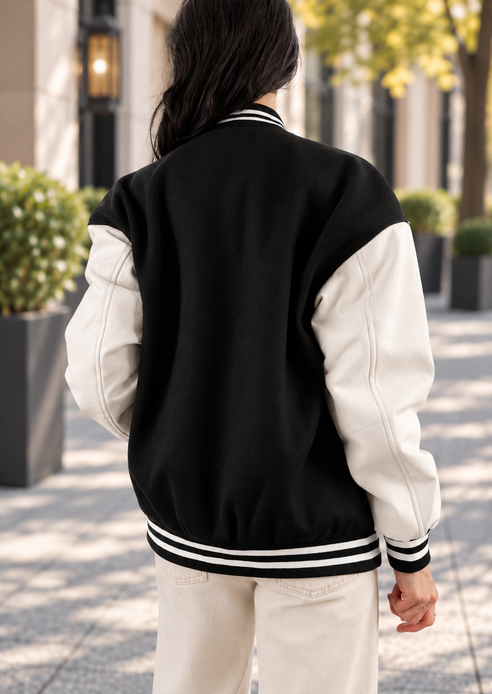 A person viewed from behind wearing a black and off-white varsity jacket with striped ribbing, standing outdoors on a paved walkway.