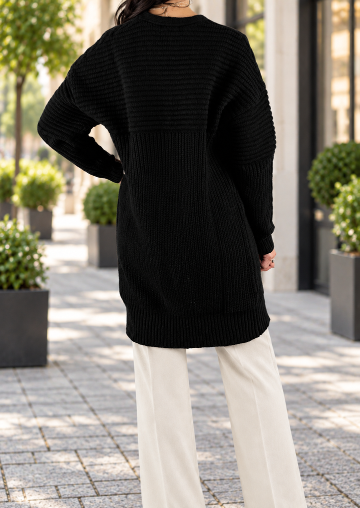 A person shown from the back wearing a black ribbed knit sweater and off-white wide-leg pants, standing outdoors on a paved walkway with potted plants and buildings in the background.
