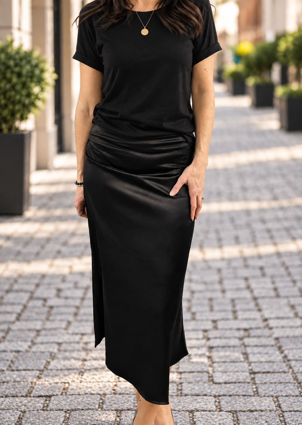 A woman wearing a black short-sleeve top and a black satin-like midi skirt with ruching, standing on a cobblestone street with blurred greenery in the background.