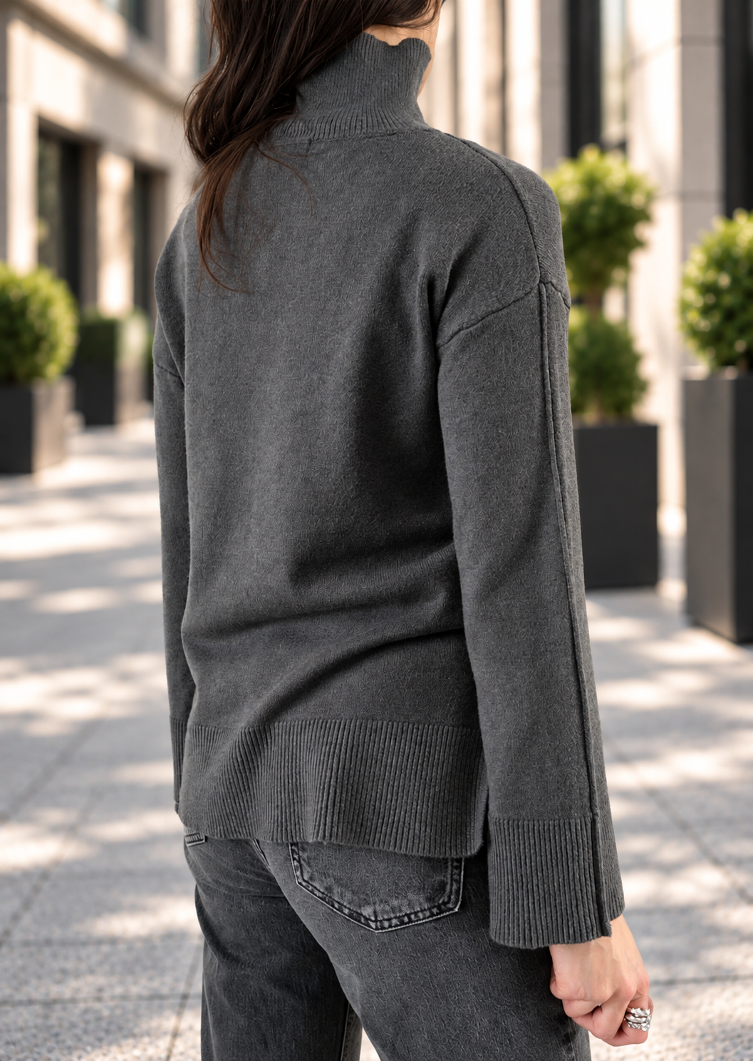 A woman wearing a dark gray ribbed mock neck sweater, shown from the back in an outdoor urban setting with buildings and potted plants in the background.