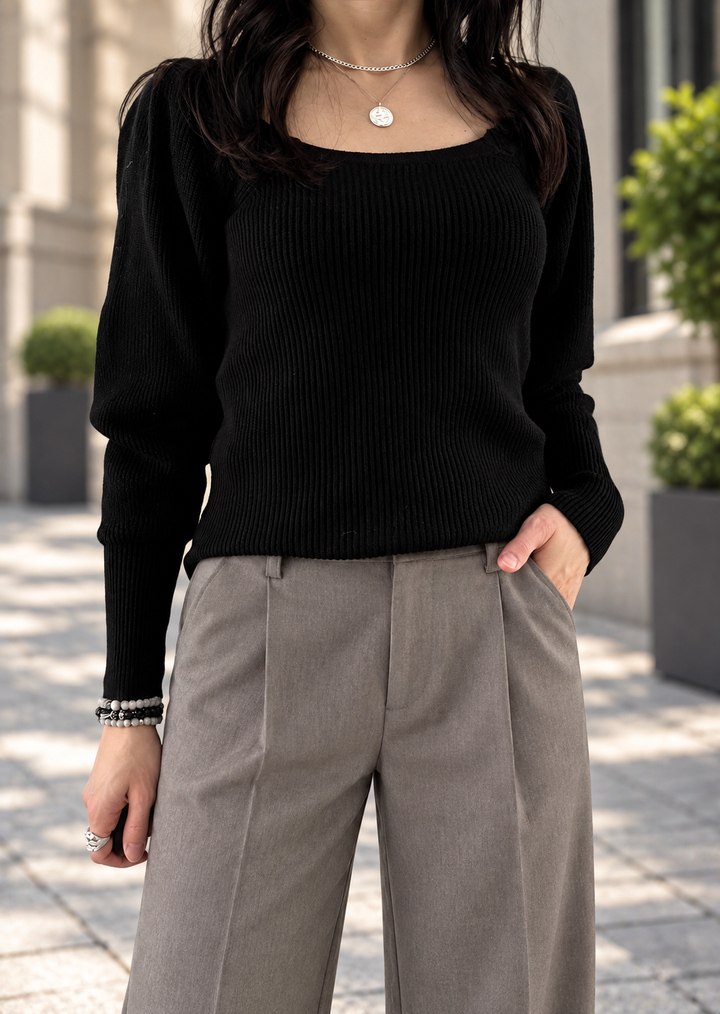 A woman wearing a black ribbed knit long-sleeve sweater with a wide scoop neckline and pleated taupe-gray wide-leg trousers, standing outdoors on a paved walkway.