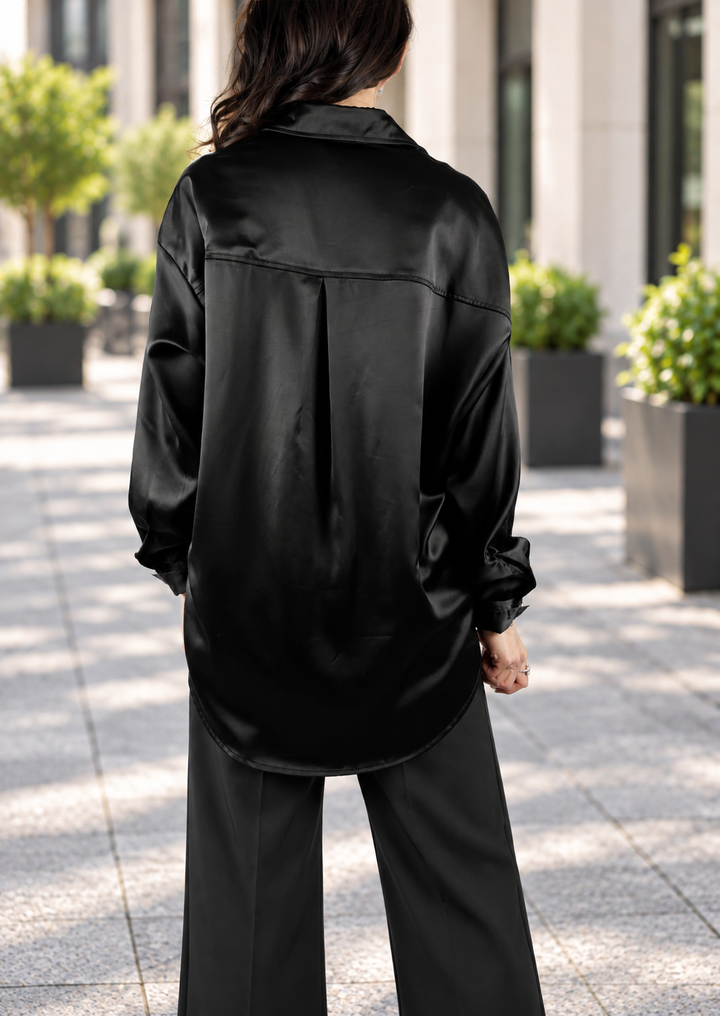 A woman viewed from behind wearing a black satin button-up shirt and black wide-leg trousers, standing on a paved outdoor walkway with blurred greenery and building columns in the background.