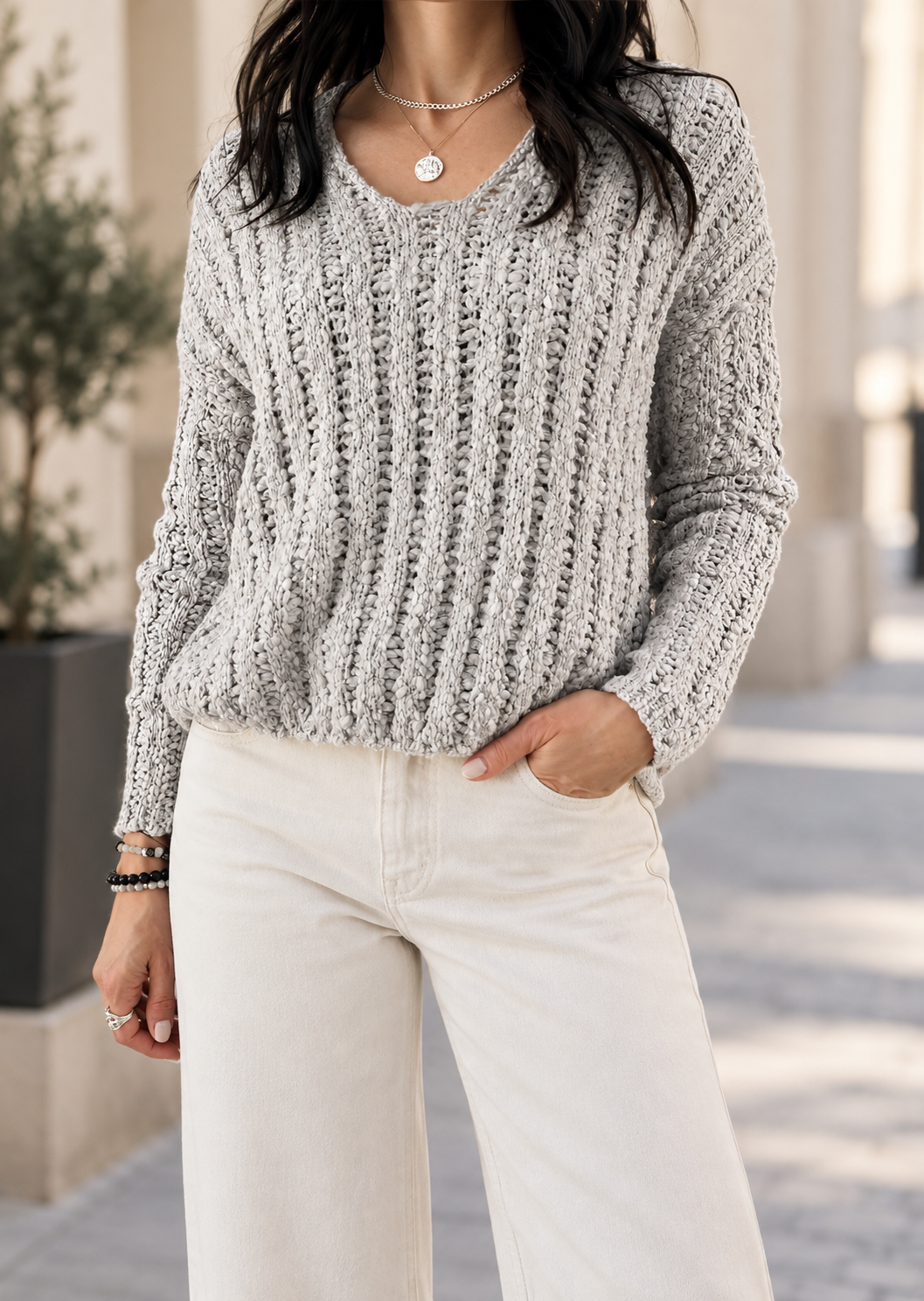 A woman wearing a light gray marled cable-knit sweater and off-white wide-leg pants, standing outdoors in a sunny urban setting.
